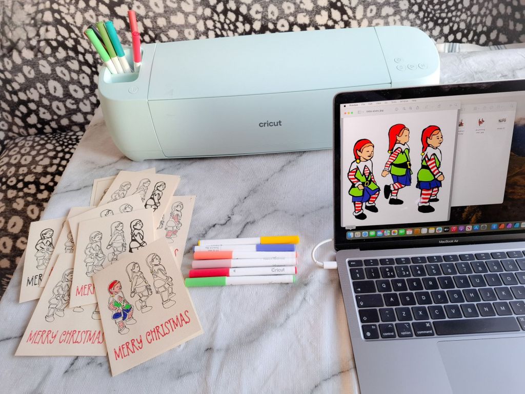 Computer screen showing the Little Elves design, plus a Cricut Explore 3 and a batch of homemade cards in various stages of being inked and coloured in